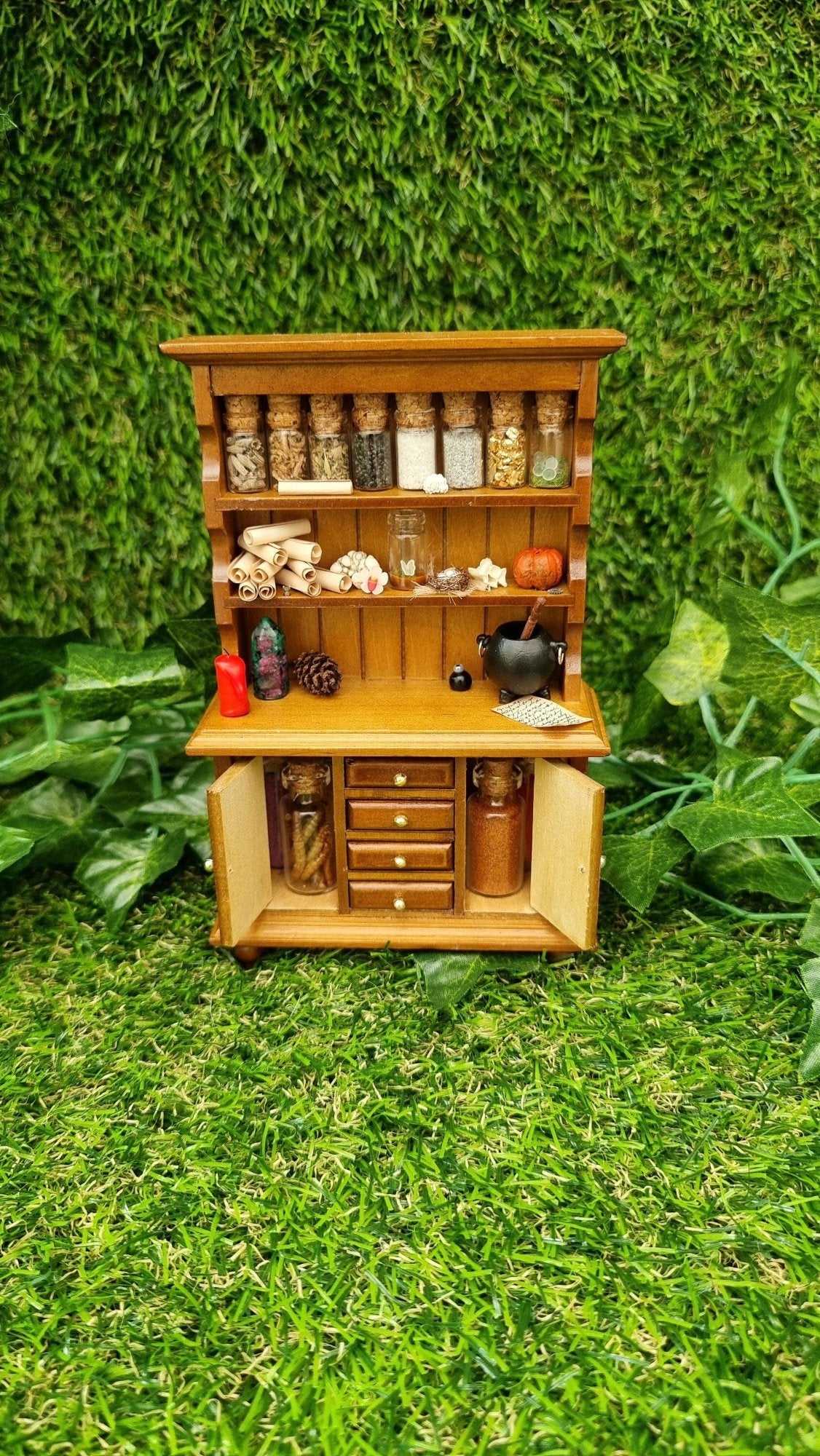 A 1:12 miniature brown dresser is shown against a green background. The dresser has 2 shelves with filled bottles, parchment rolls and curios. The counter top has a candle and cauldron. There are 2 open cupboard doors with more bottles and 4 drawers