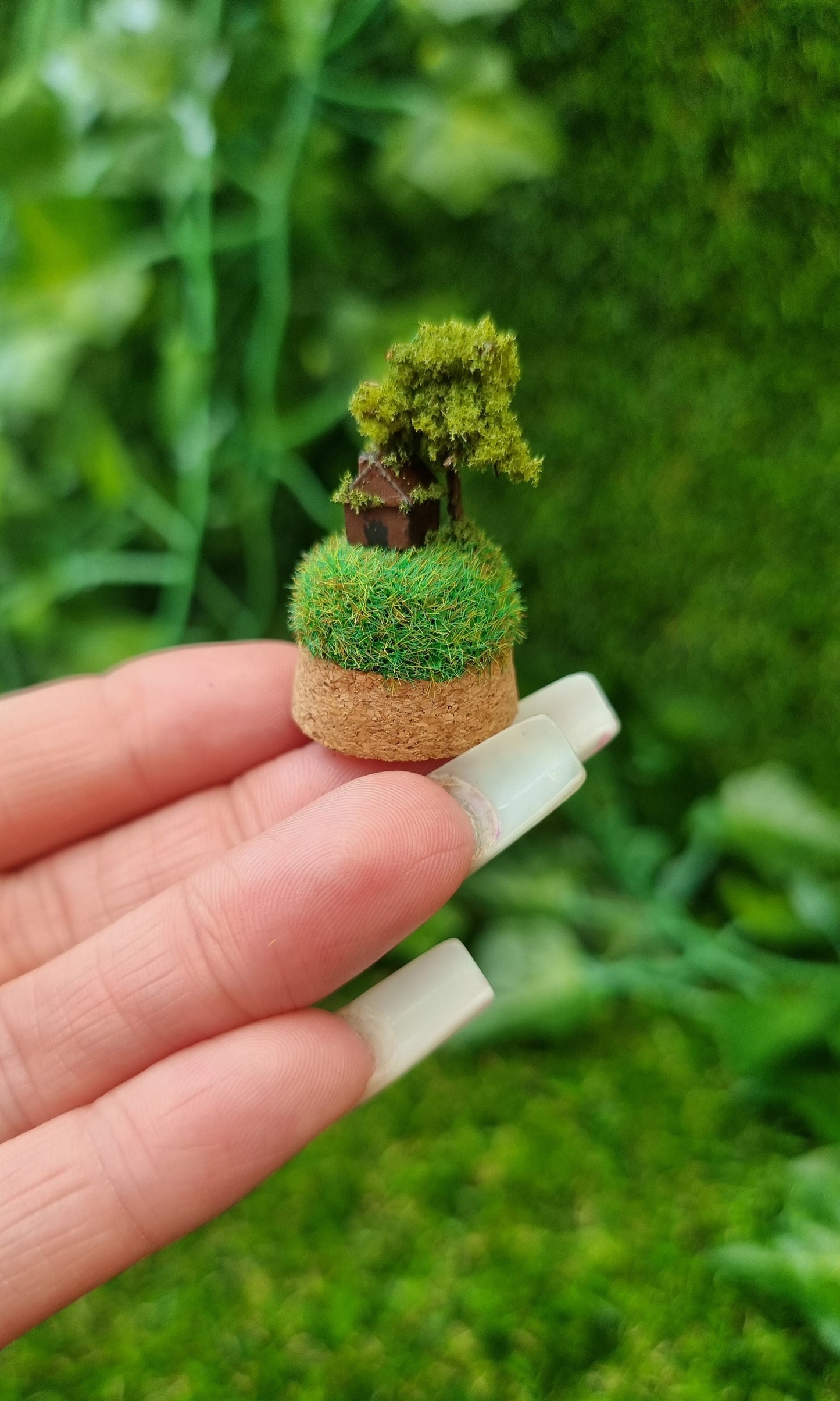 Miniature cork base containing a tiny brown house and a tree on top a grassy hill sits on a hand