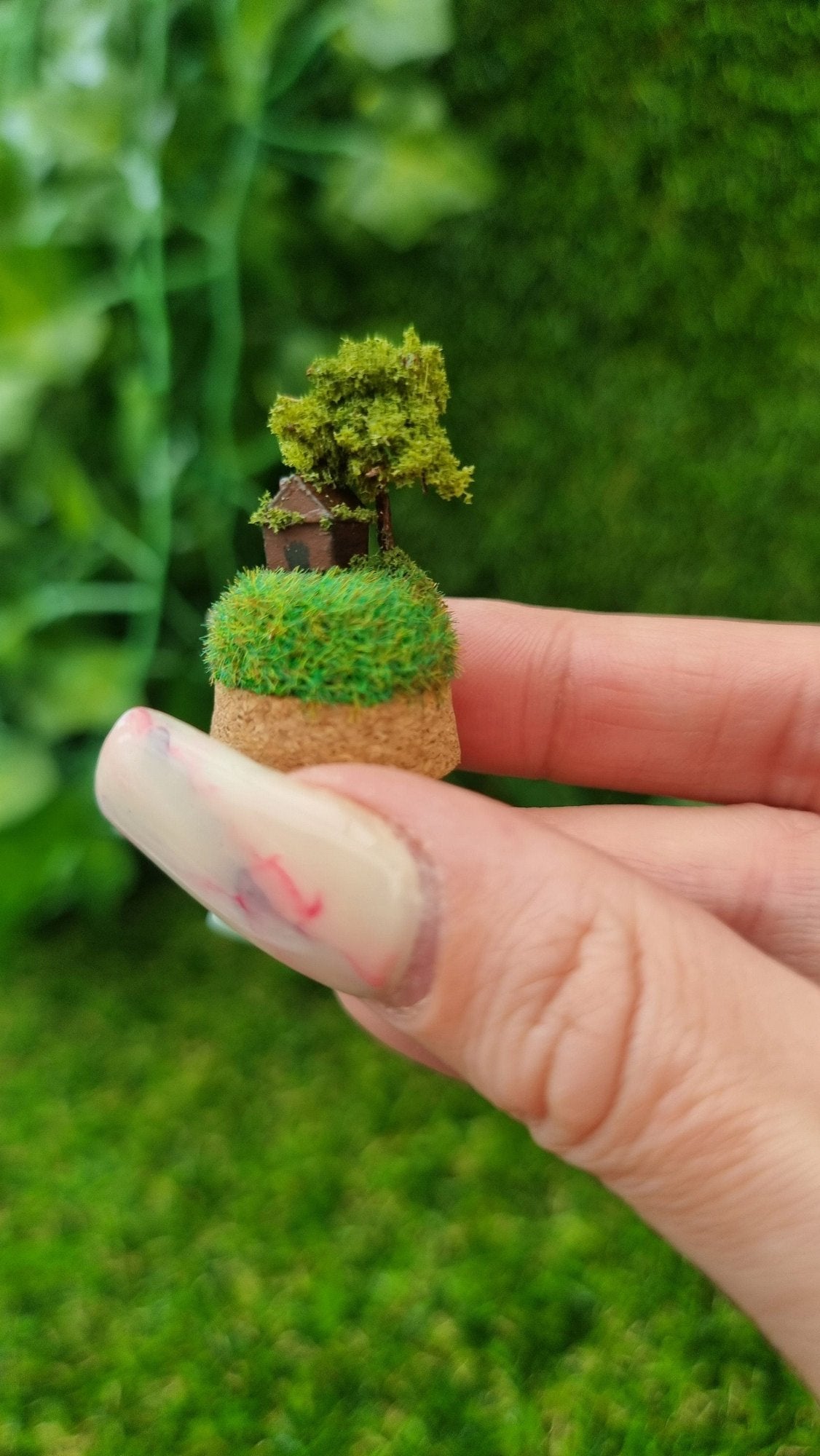 Miniature diorama on a cork base. It has a tiny brown house and a tree on top of a green grassy hill, sitting on a hand.