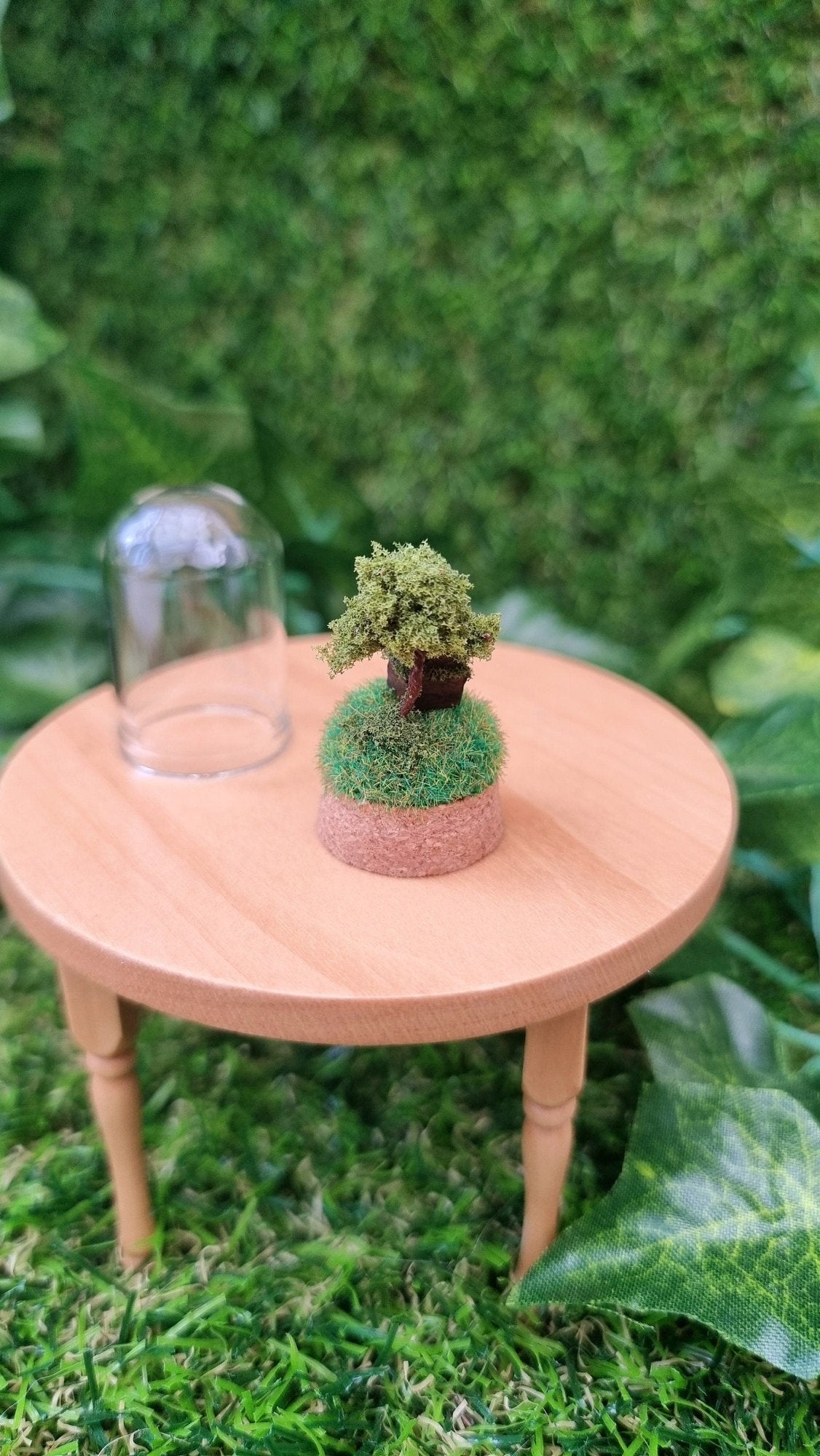 Back view of a miniature diorama on a cork base. It has a tiny brown house and a tree on top of a grassy hill, sitting on a 1:12 scale table. There is a small glass bell jar lid sat behind it