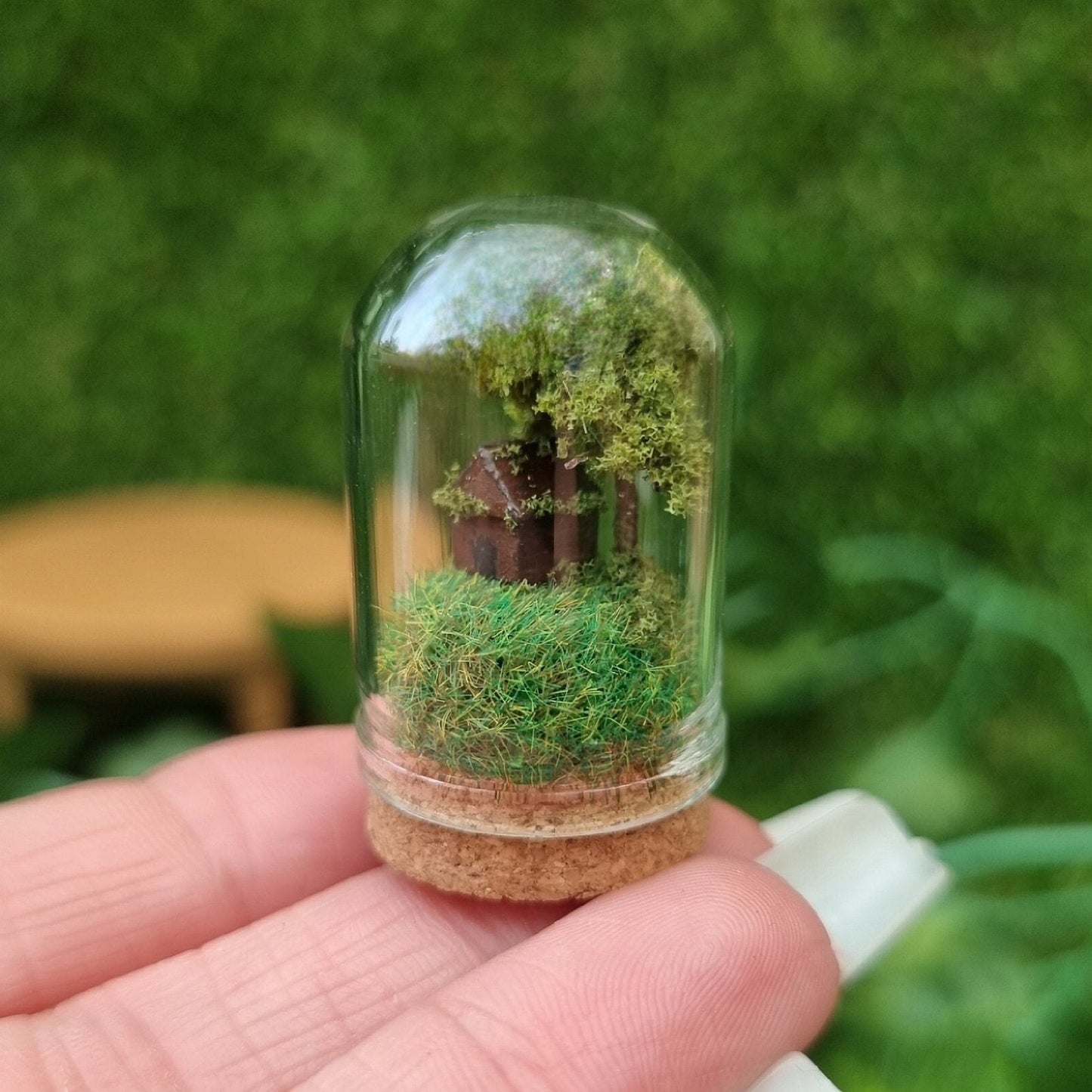 Miniature glass bell jar with cork base contzining a tiny brown house and a tree on a grassy hill sits on a hand