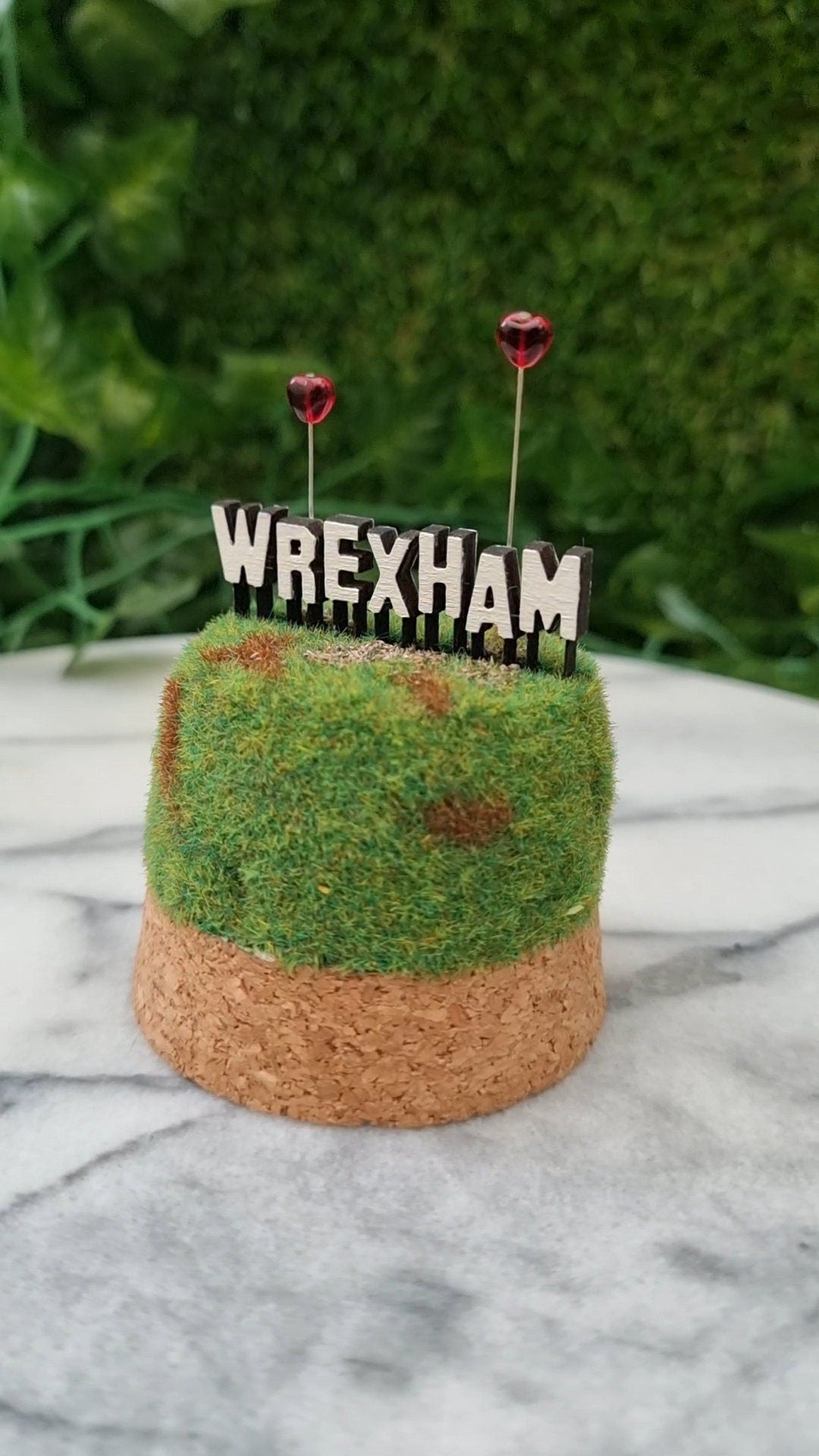 A small diorama is sat on a marble background. There is a green and brown grassy hill topped with a Hollywood-style Wrexham sign on top seen from the back. The R and A have red heart shaped balloons on wires floating above the sign