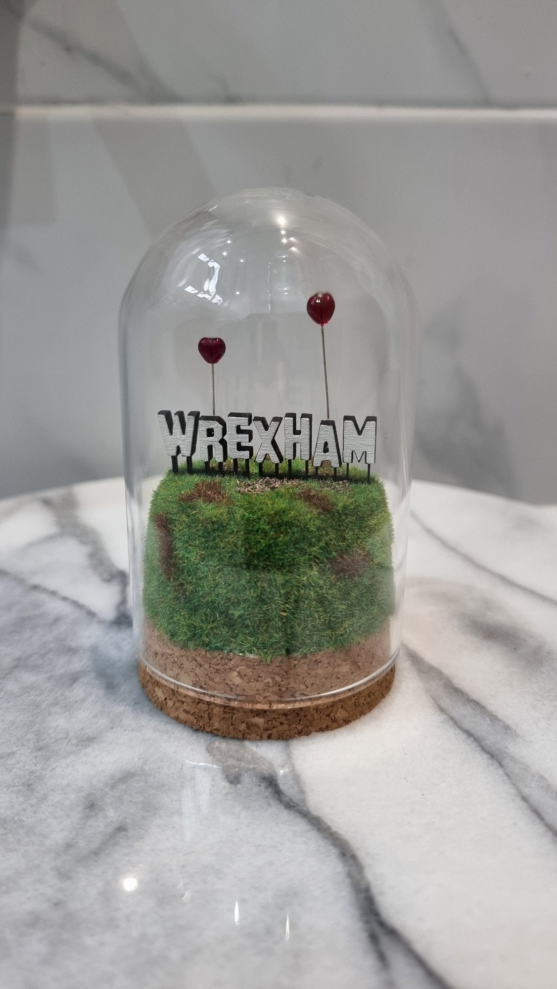 A small diorama with a glass dome cover contains a green and brown grassy hill topped with a white Hollywood-style Wrexham sign on top. The letters R and A have red heart shaped balloons on wires floating above the sign. Background or marble