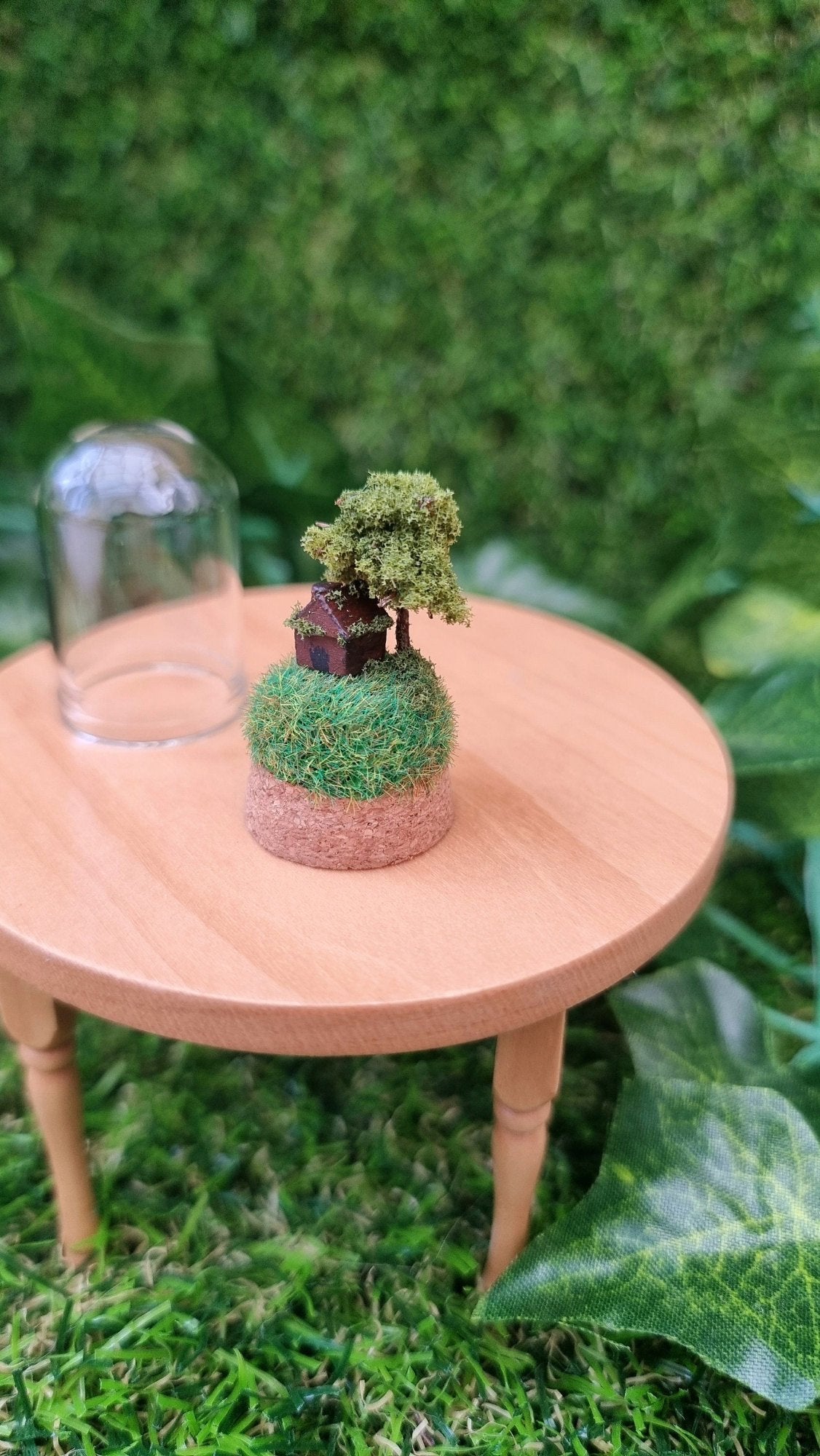 Miniature cork base containing a tiny brown house and a tree on top a grassy hill sits on a 1:12 scale table. There is a small glass bell jar lid sat behind it