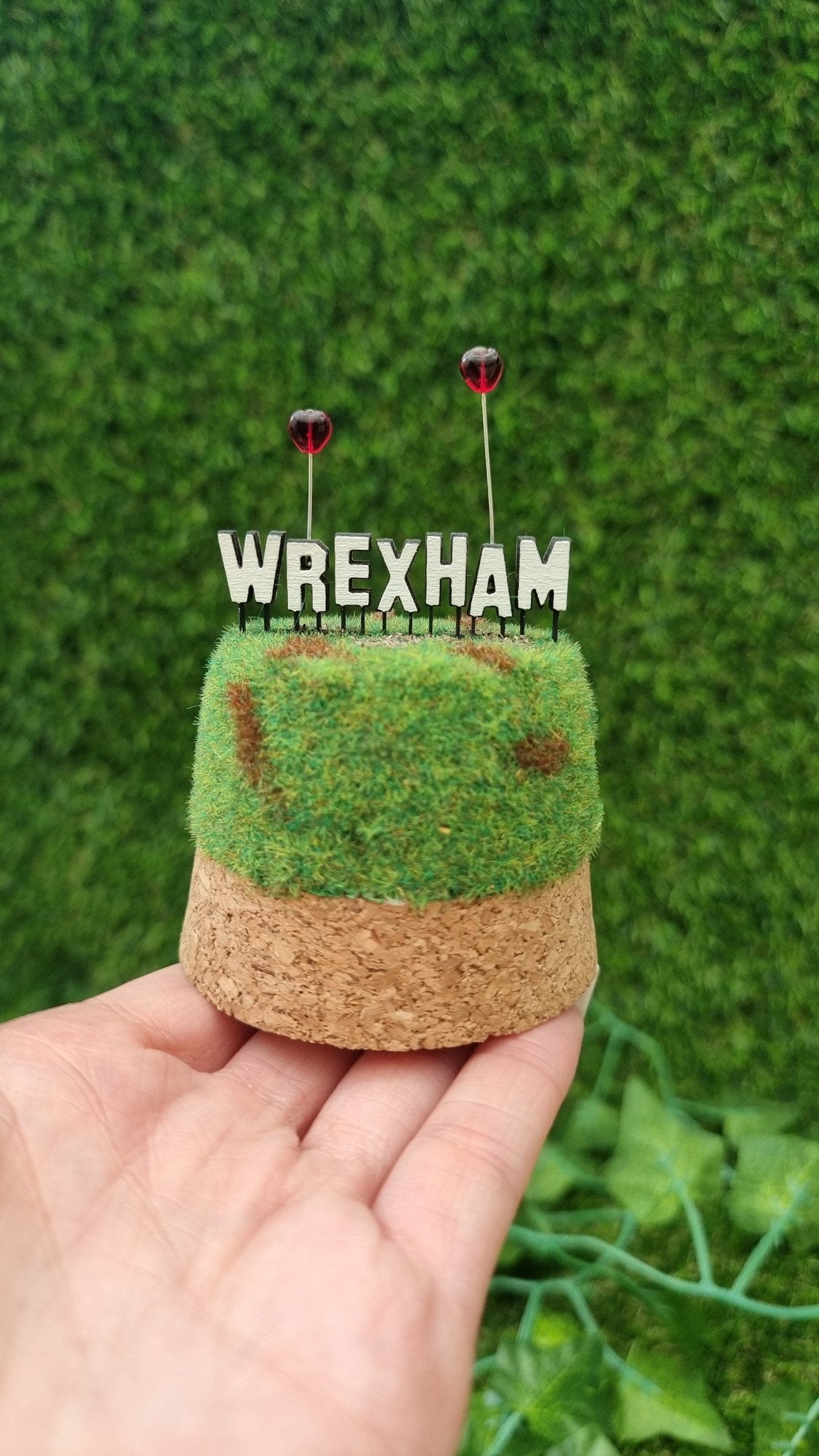 A small diorama is held on a palm. There is a green and brown grassy hill topped with a Hollywood-style Wrexham sign on top. The R and A have red heart shaped balloons on wires floating above the sign