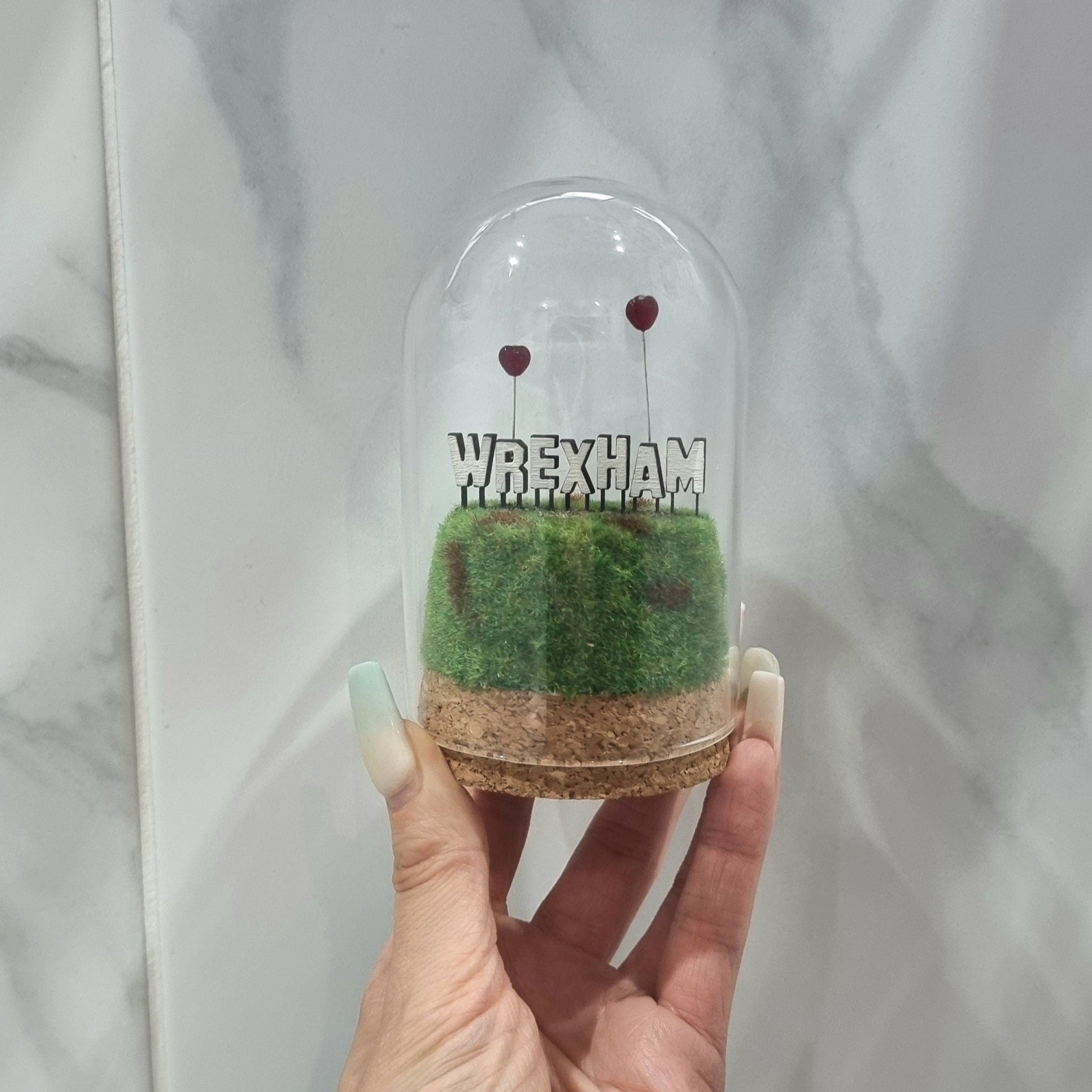 A small diorama with a glass dome cover is held by a hand. There is a green and brown grassy hill topped with a white Hollywood-style Wrexham sign on top. The letters R and A have red heart shaped balloons on wires floating above the sign.