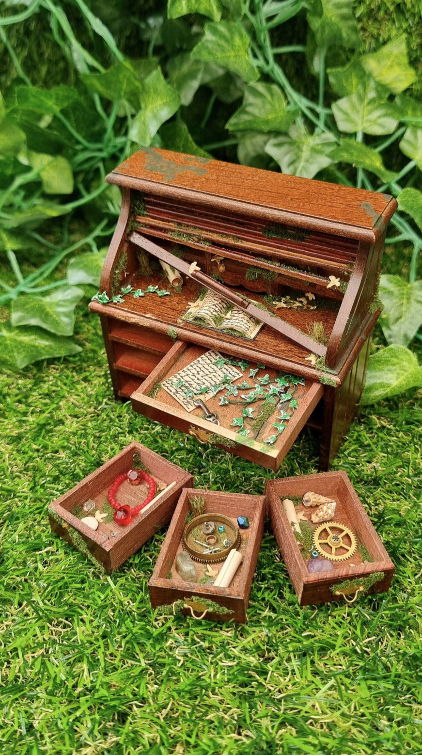 1:12 scale miniature abandoned rolltop desk drawer has an open drawer revealing moss and ivy. Three other drawers sit in front, with paper, dead leaves, trinkets , cogs and gemstones. Green ivy creeps across the desk top towards paper and a mossy book