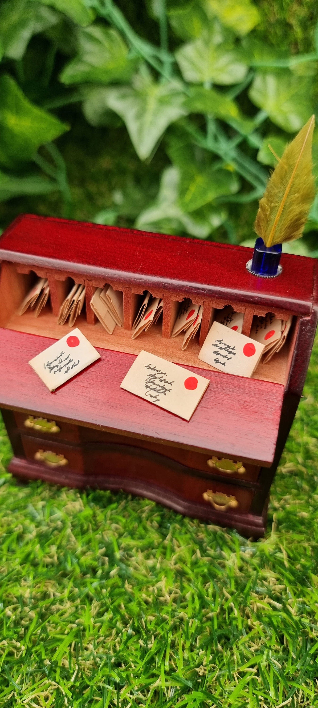 A 1:12 scale brown miniature bureau desk sits against a green background. The desk is topped with an inkwell and quill. The bureau desk top is open, with 3 letters out, and lots of tiny letters sat inside little pigeon holes