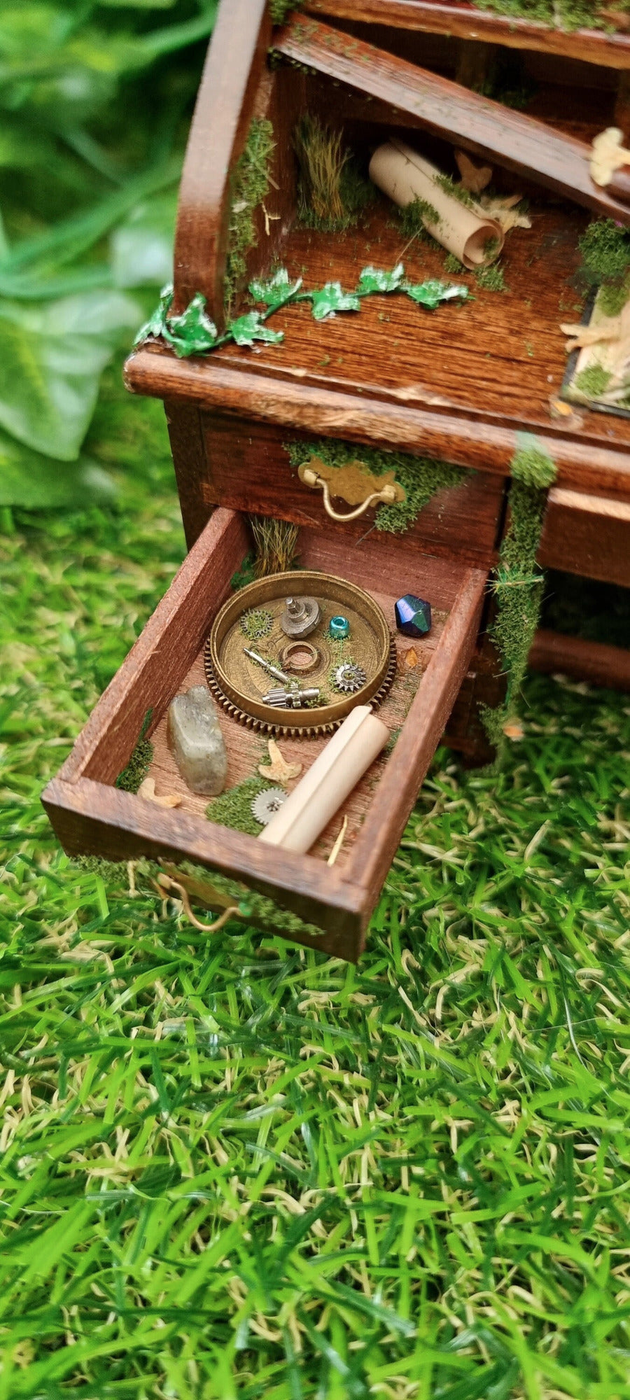 1:12 scale miniature abandoned desk drawer is open, revealing moss, grass, scroll of paper, dead leaves, trinkets and a gemstone. Green ivy creeps across the desk top towards a roll of paper and a mossy book