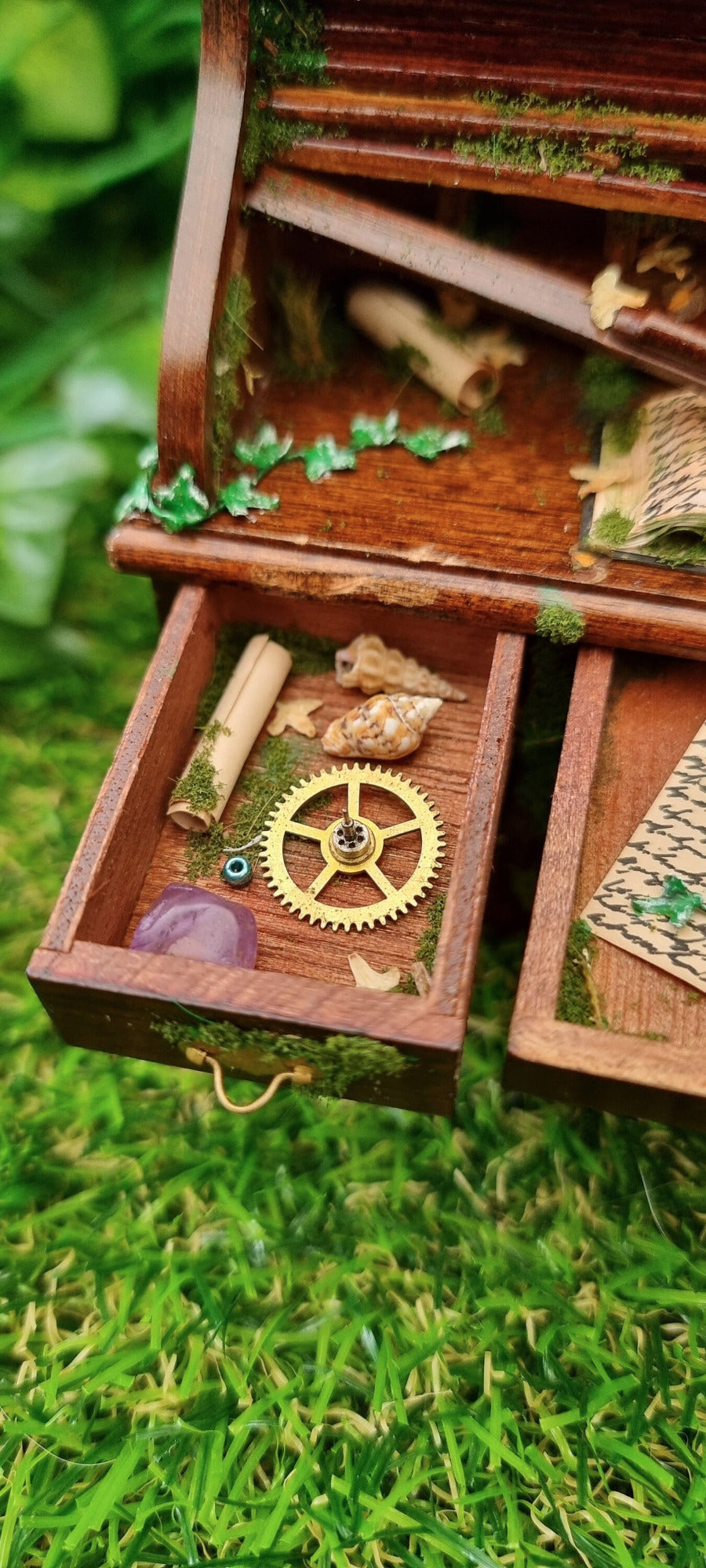 1:12 scale miniature abandoned desk drawer is open, revealing moss, cog, gemstone, shells and dead leaves. Green ivy creeps across the desk top towards a moss piece of paper and book