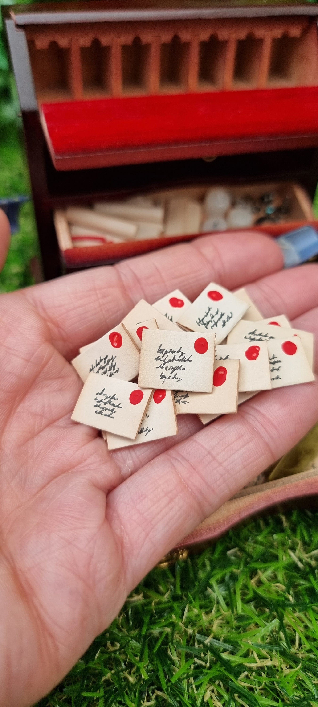 A hand holds a stack of tiny 1:12 scale aged letters, each with a tiny address and red stamp on the front. The open miniature desk sits in the background