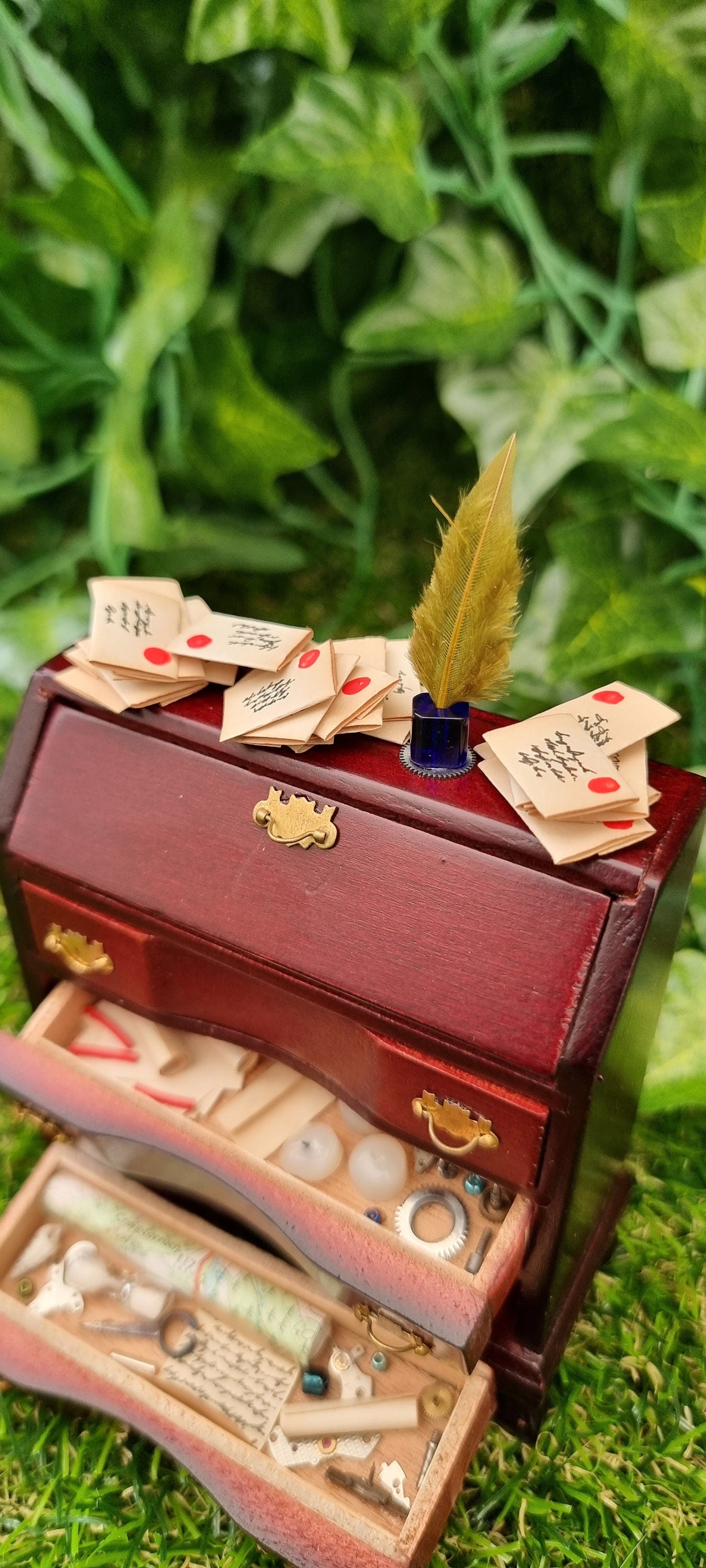 A 1:12 scalebrown miniature bureau desk sits against a green background. The desk is topped with stacks of old letters, inkwell and quill. Two drawers are open, revealing papers, candles, map, curios ad trinkets