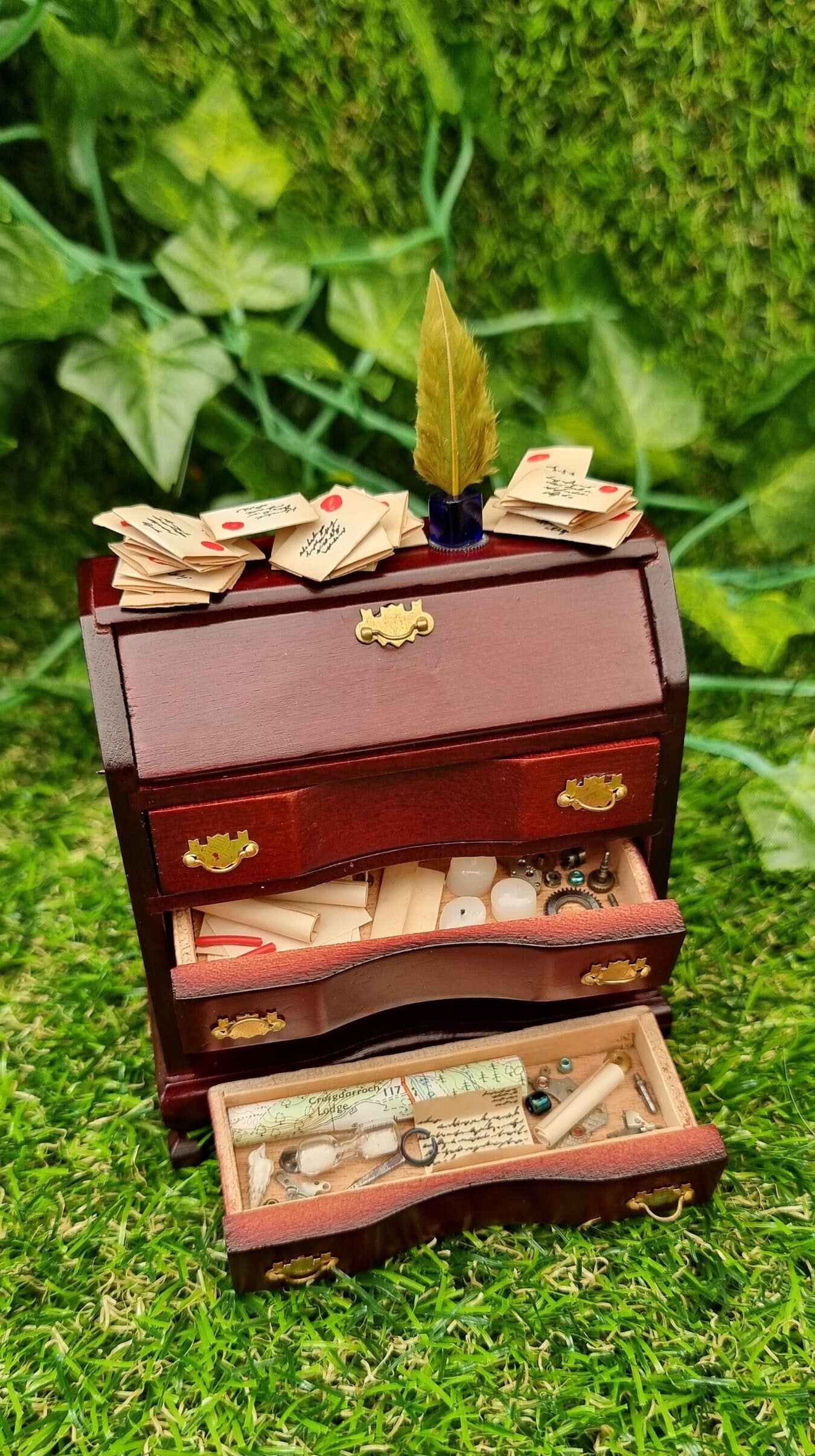 A 1:12 scale brown miniature bureau desk sits against a green background. The desk is topped with stacks of old letters, inkwell and quill. Two drawers are open, revealing papers, candles, map, curios ad trinkets.