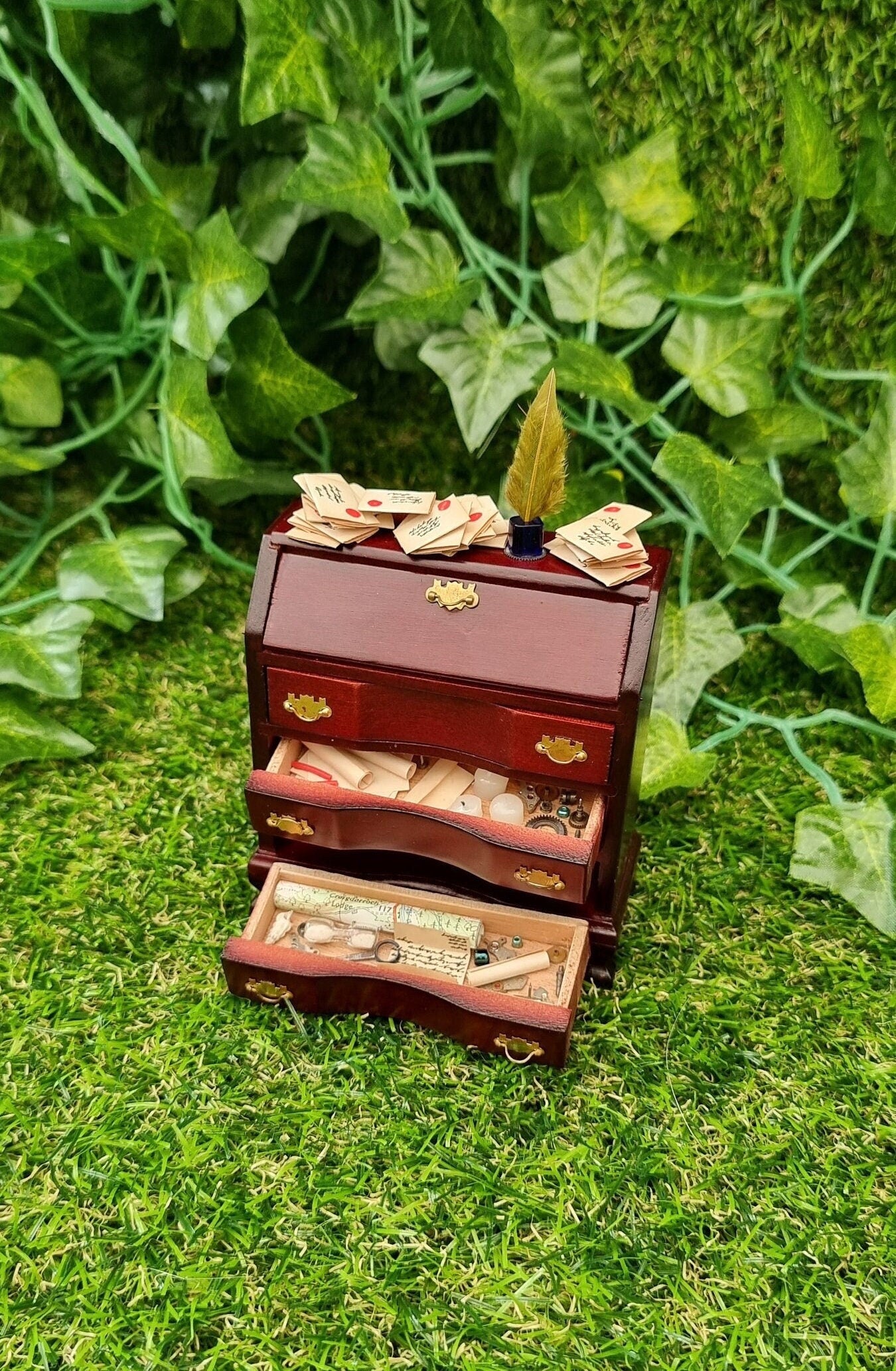 A 1:12 scale brown miniature bureau desk sits against a green background. The desk is topped with stacks of old letters, inkwell and quill. Two drawers are open, revealing papers, candles, map, curios and trinkets