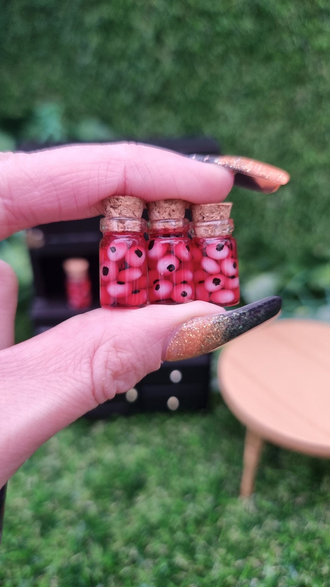 Two fingers hold 3 1:12 scale miniature glass jars containing eyeballs and red liquid. The miniature jars are topped with cork stoppers