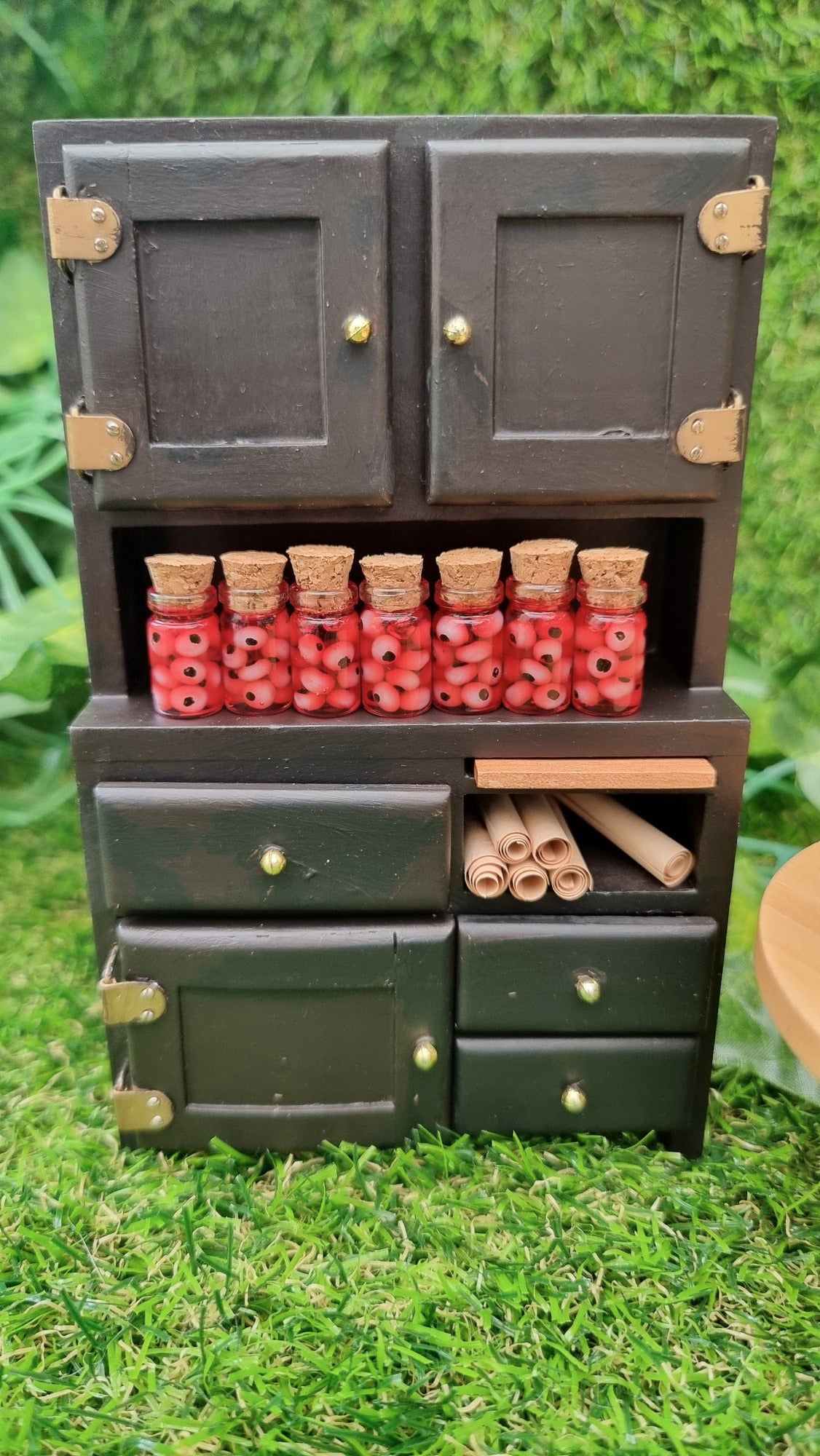 A 1:12 miniature black dresser holds 7 miniature glass jars containing eyeballs and red liquid.  The miniature jars are topped with cork stoppers