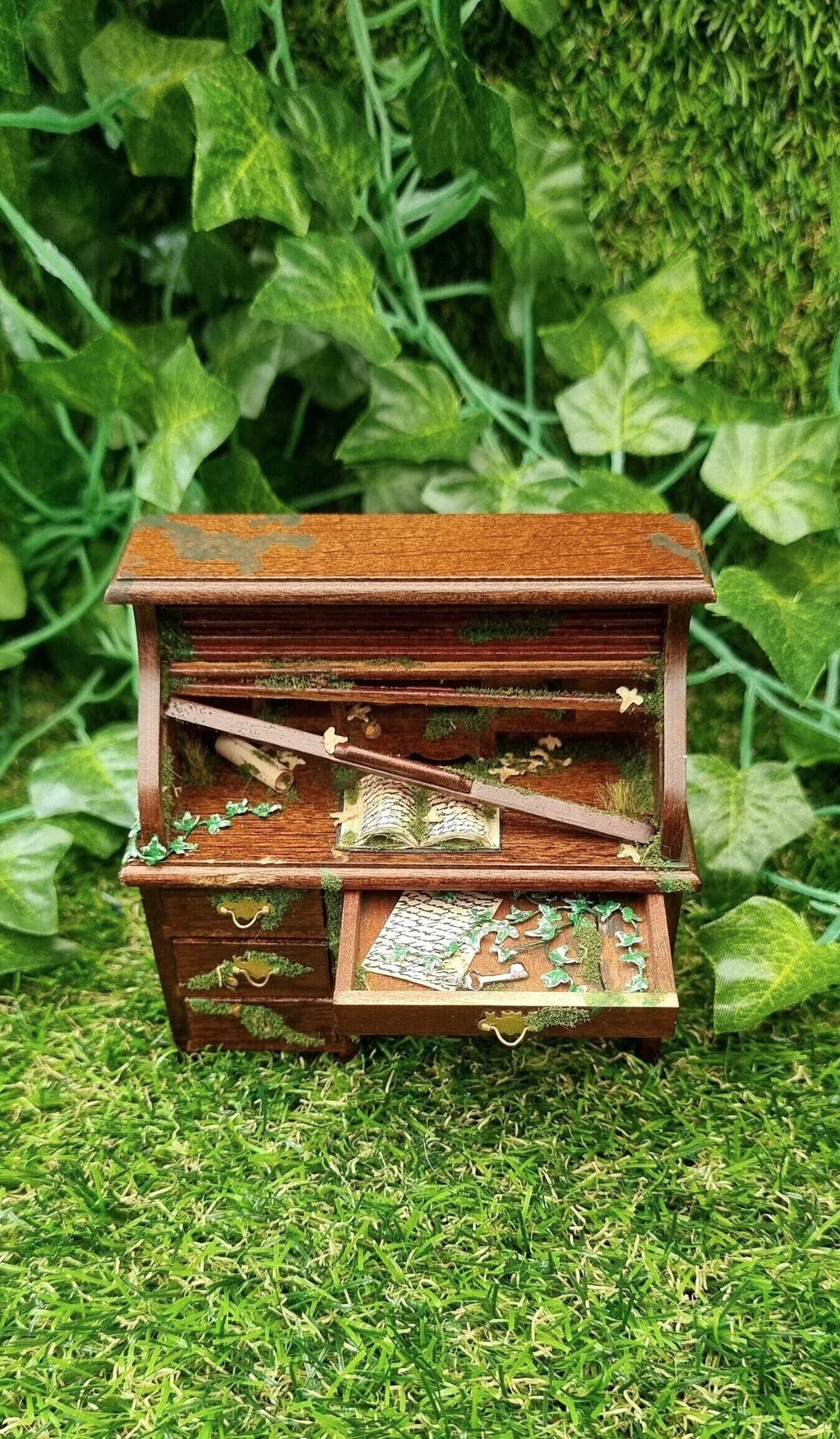 1:12 scale miniature abandoned rolltop desk with the top drawer open.  The drawer contains green creeping ivy, moss, an old letter and a silver key. The desk top has a mossy old book, leaves and a broken wooden slat of the roll top