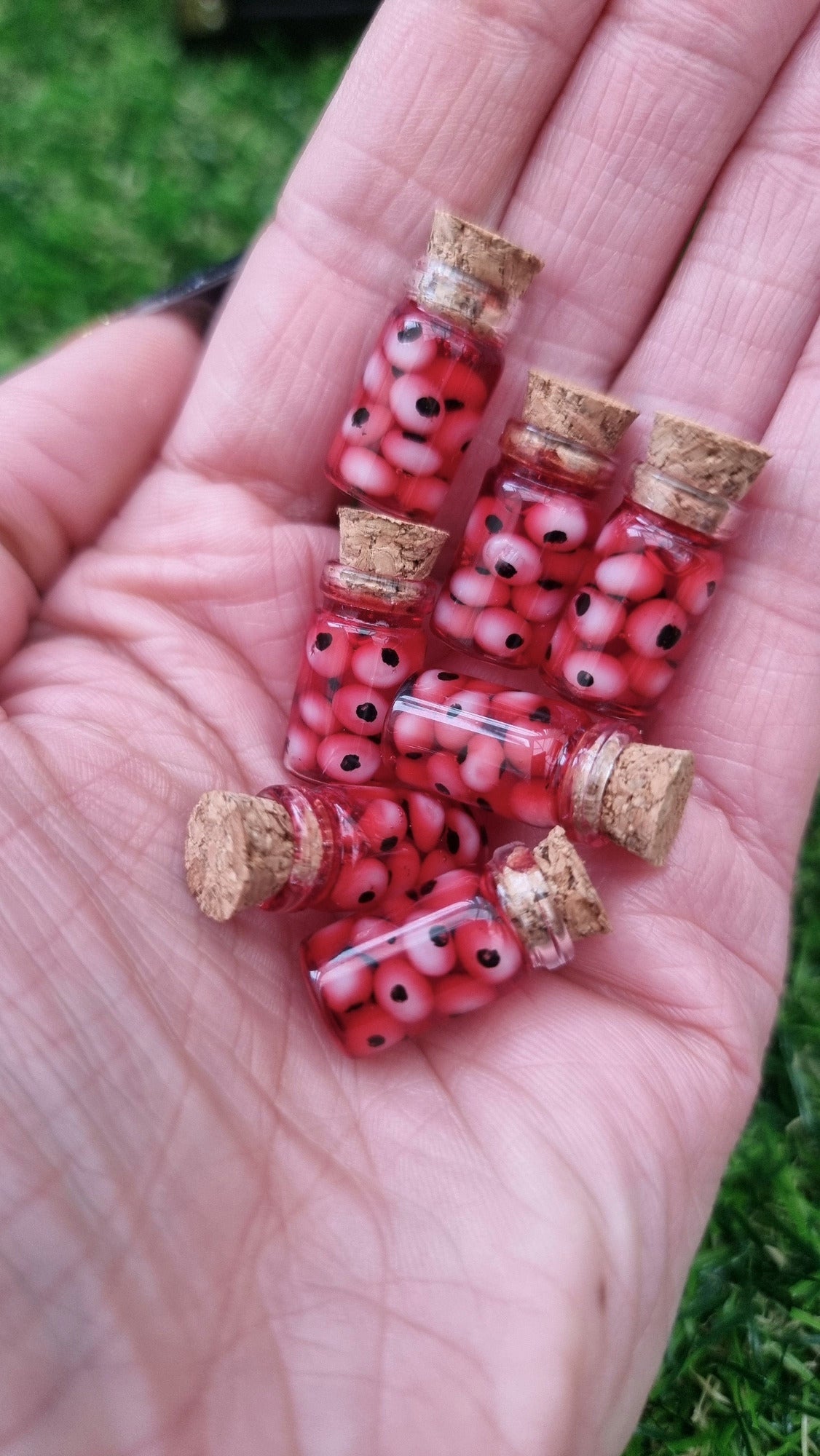 A hand holds 7 1:12 scale miniature glass jars containing eyeballs and red liquid.  The miniature jars are topped with cork stoppers