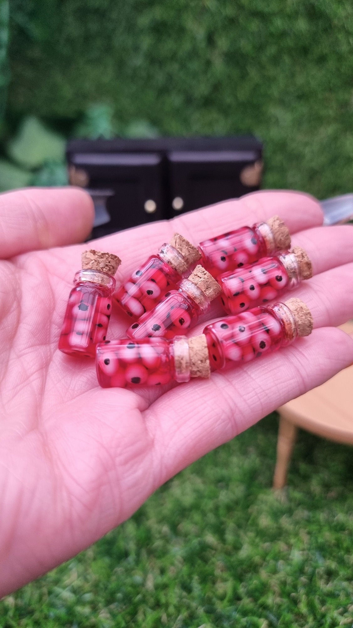 A hand holds 7 1:12 scale miniature glass jars containing eyeballs in a red liquid.  The miniature jars are topped with cork stoppers