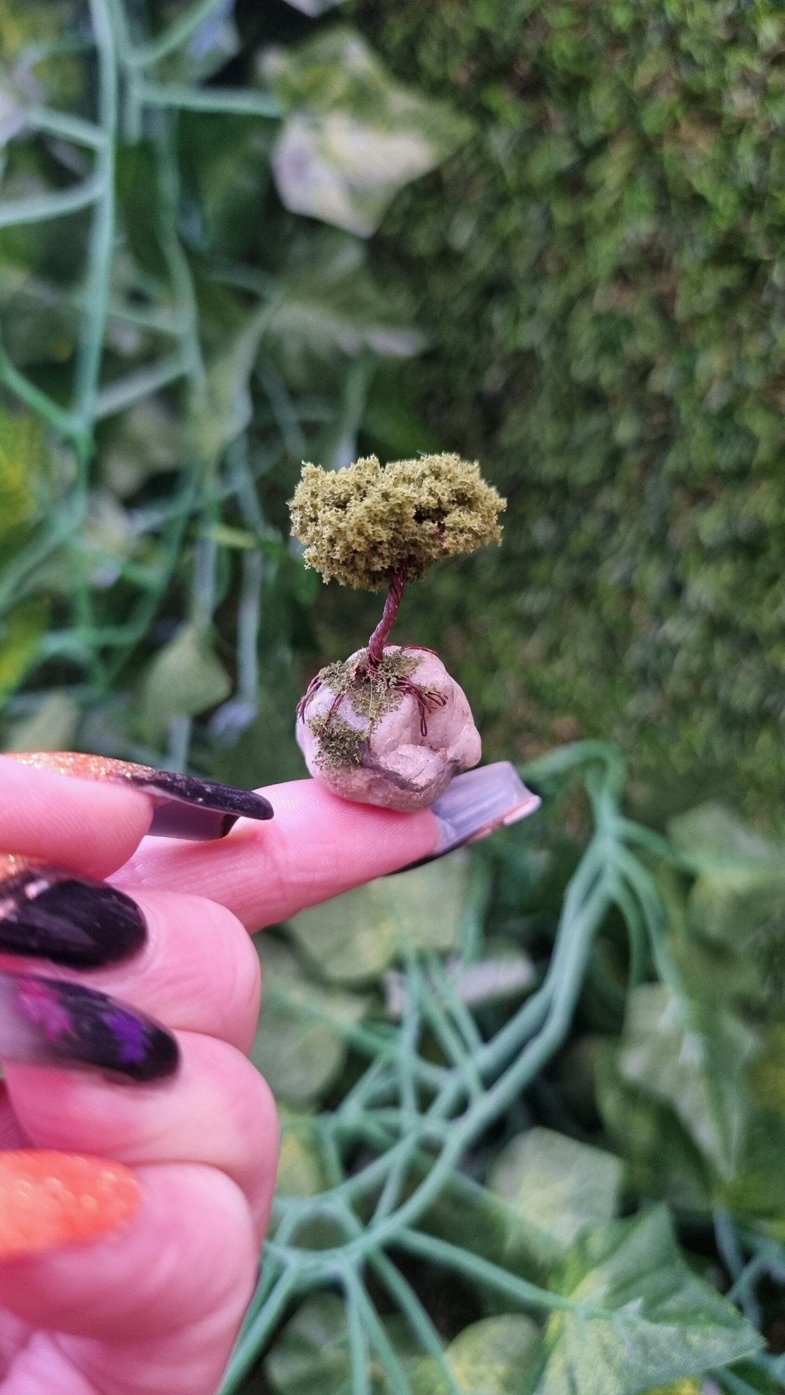 A 1:12 scale bonsai tree is held on the end of a finger. The bonsai is mounted on a quartz stone with roots and moss creeping down