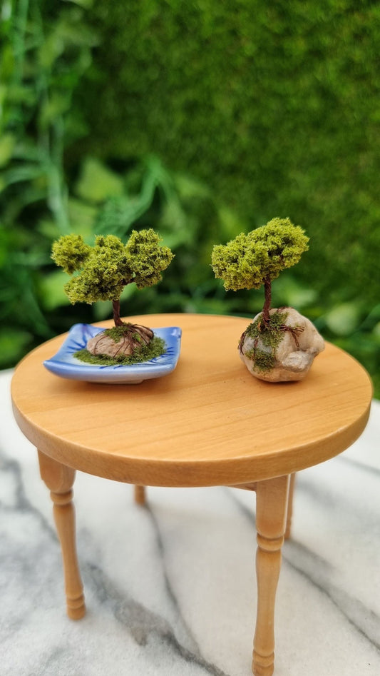 2 1:12 scale bonsai trees sit on a miniature table. One bonsai is mounted on a quartz stone and the other bonsai is mounted on a small stone on a blue and white square plate