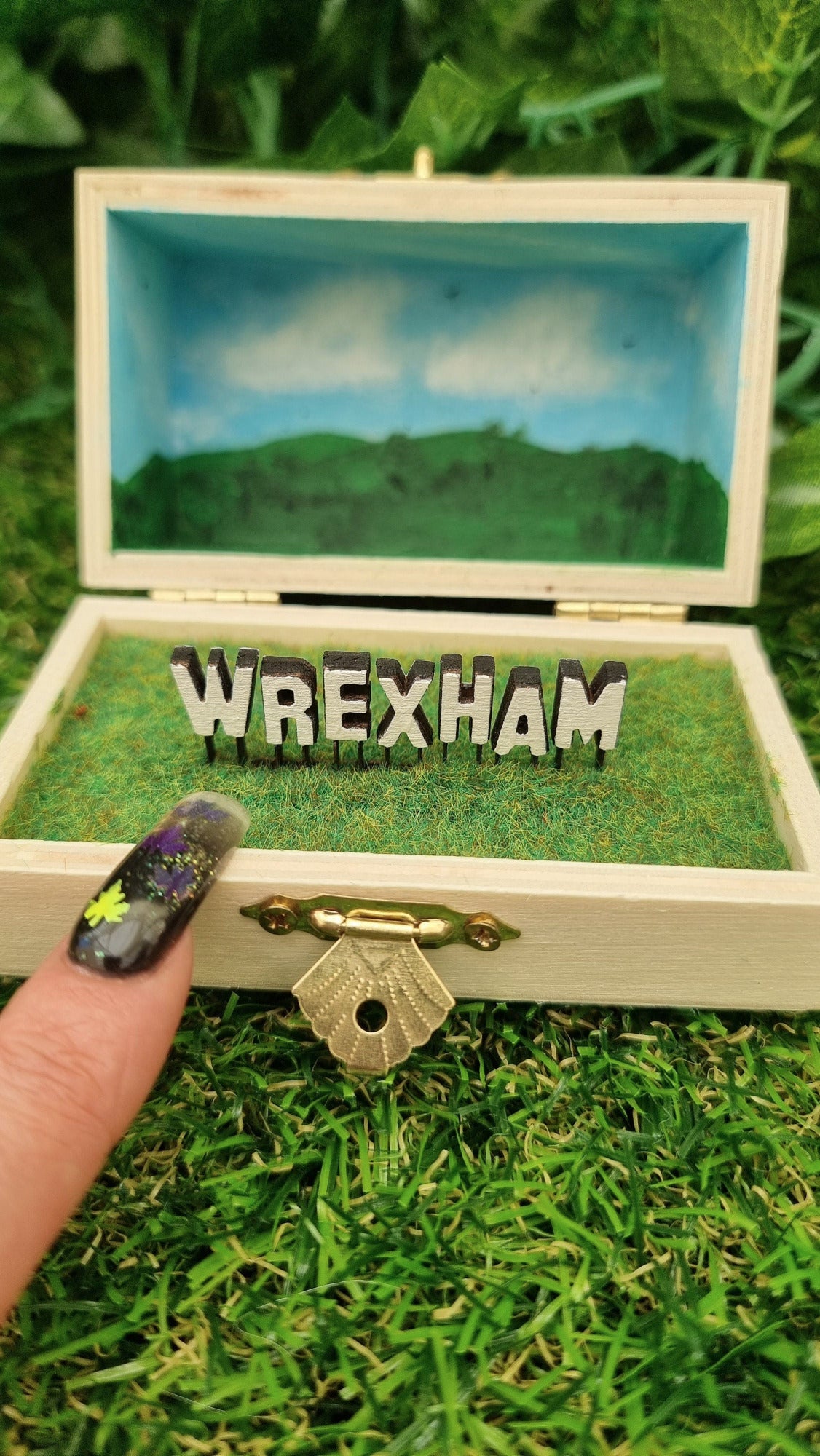 A small hinged wooden box sits open. The box contains a white hollywood-style Wrexham sign on flocked grass field. The deep lid of the box sits behind with a painted countryside scene with green hills, blue sky, white clouds and soaring birds