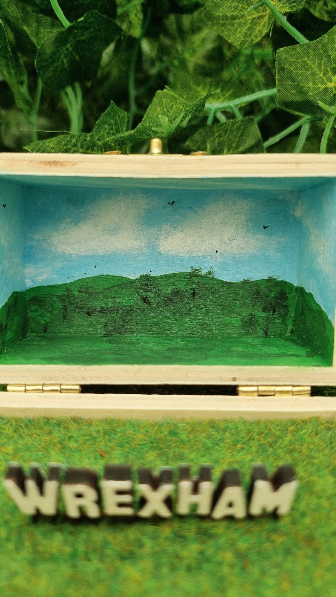 Details of a small open hinged wooden box diorama. The deep lid of the box has a painted countryside scene with green hills, blue sky, white clouds and soaring birds. A hollywood style Wrexham sign sits in front