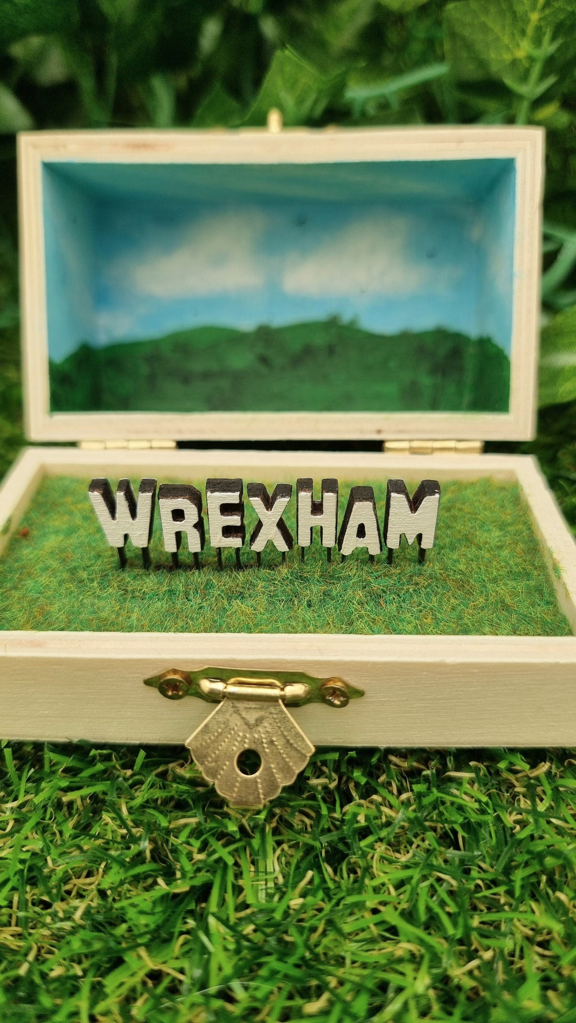 A small hinged wooden box sits open. The box contains a white hollywood-style Wrexham sign on flocked grass field. The deep lid of the box sits behind with a painted countryside scene with green hills, blue sky, white clouds and soaring birds