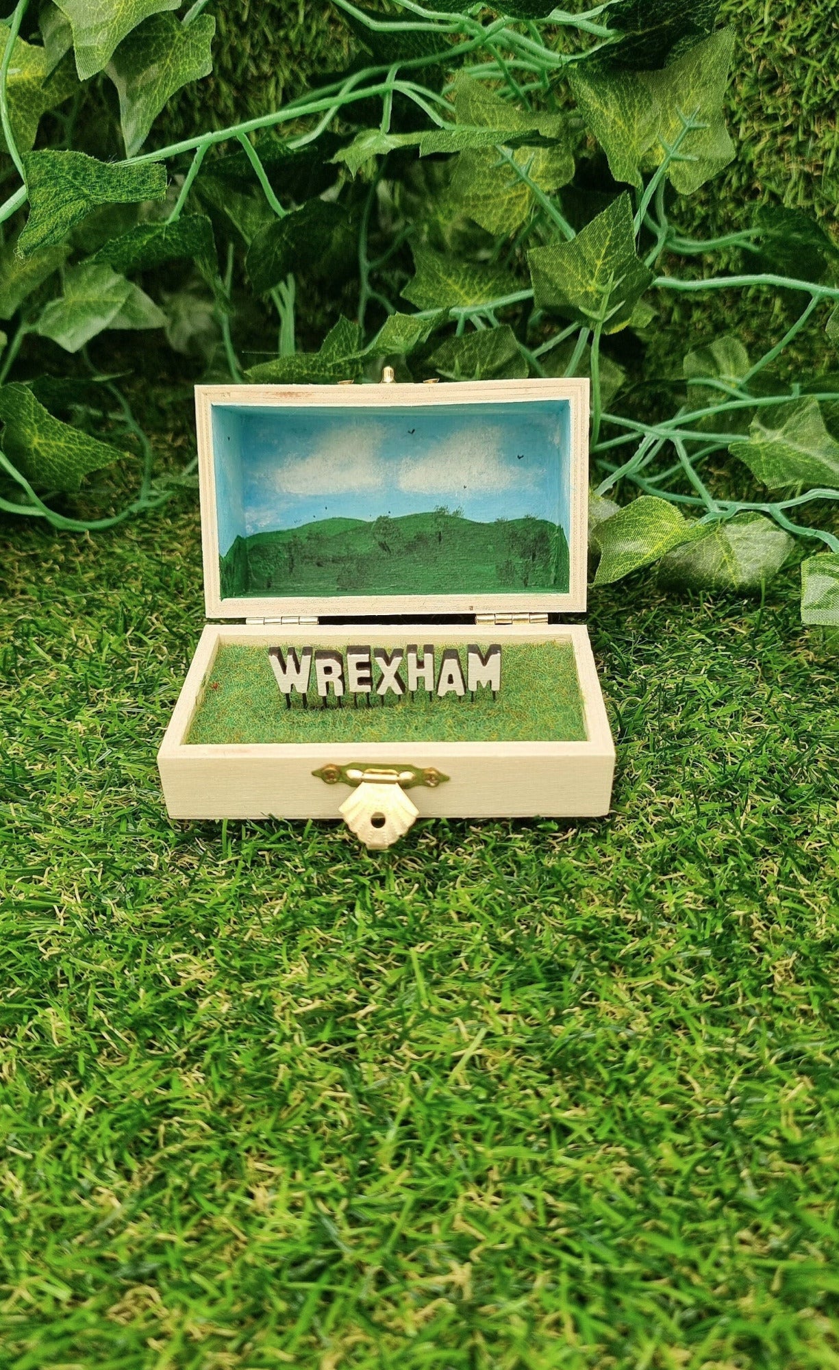 A small hinged wooden box sits open. The box contains a white hollywood-style Wrexham sign on flocked grass field. The deep lid of the box sits behind with a painted countryside scene with green hills, blue sky, white clouds and soaring birds