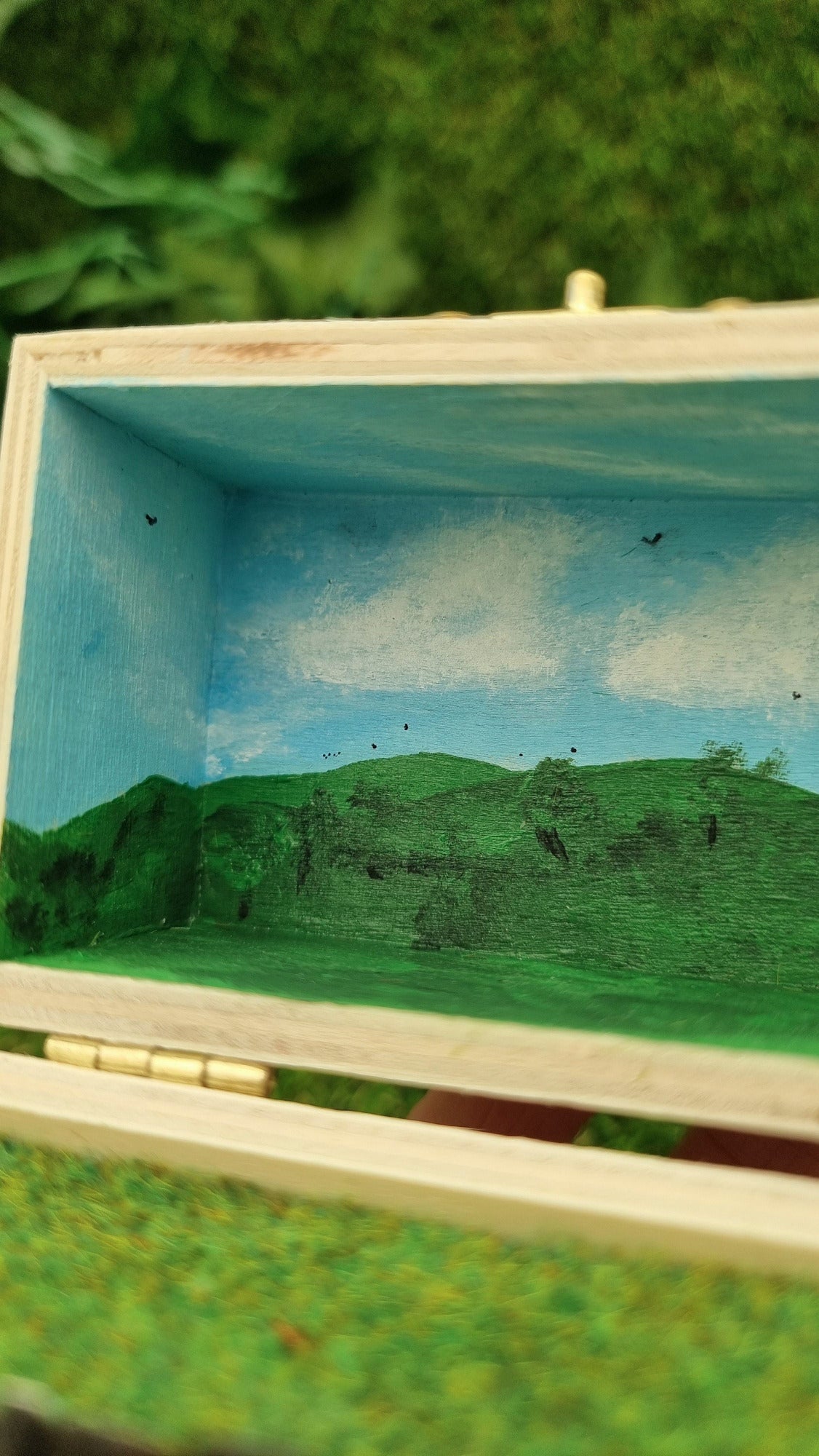 A small hinged wooden box sits open. The box contains a white hollywood-style Wrexham sign on flocked grass field. The deep lid of the box sits behind with a painted countryside scene with green hills, blue sky, white clouds and soaring birds