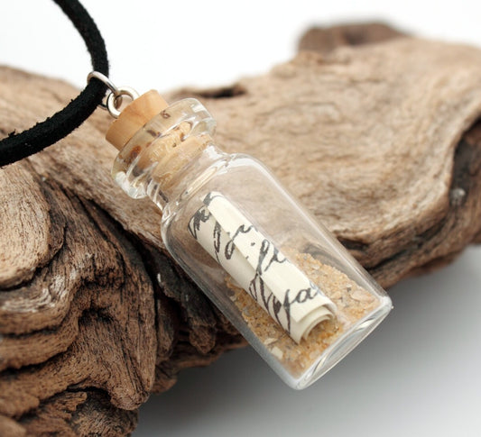 Small message in a bottle glass jar necklace containing a small scroll of paper and golden brown sand. The  jar is displayed on a black coloured cord attached to the cork lid