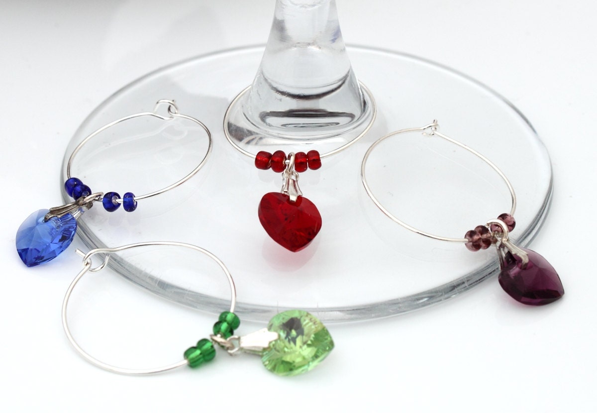 4 wine glass charms are arranged on top of a wine glass base on a white background. The wine glass charms are round wire hoops, each with different coloured seed beads and a Swarovski crystal heart in red, green, blue and lavender