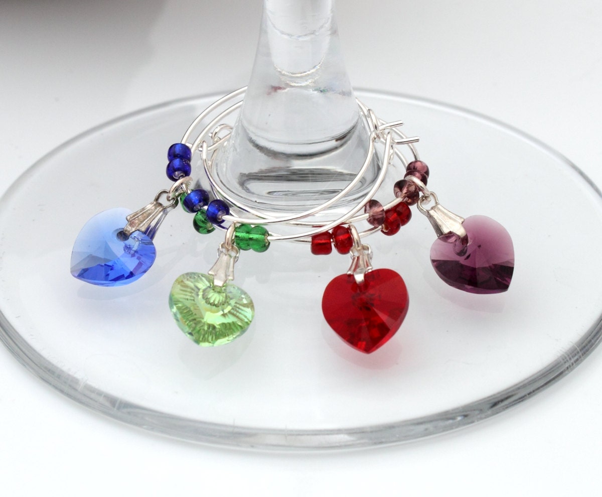 4 wine glass charms are arranged  side by side on top of a wine glass bases on a white background. The wine glass charms are round wire hoops, each with different coloured seed beads and a Swarovski crystal heart in red, green, blue and lavender